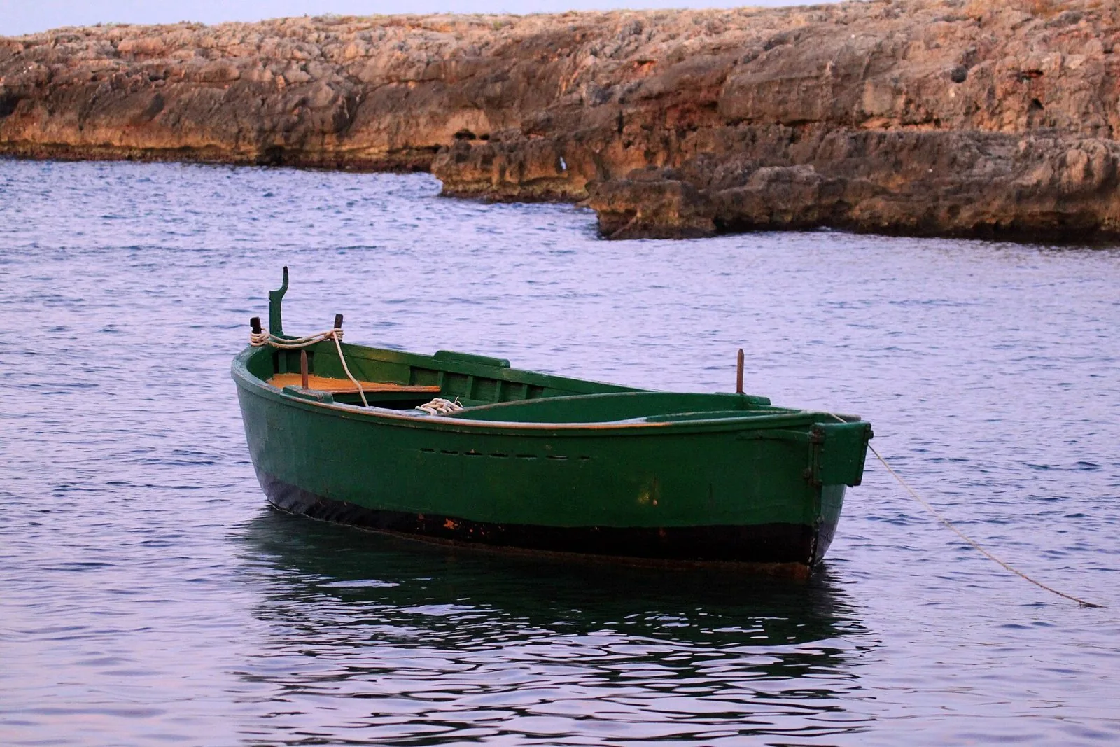 Piccola barca da pesca in legno verde ormeggiata nell'acqua calma davanti a una costa di roccia calcarea, nella luce rosata del tramonto a Cala Incina