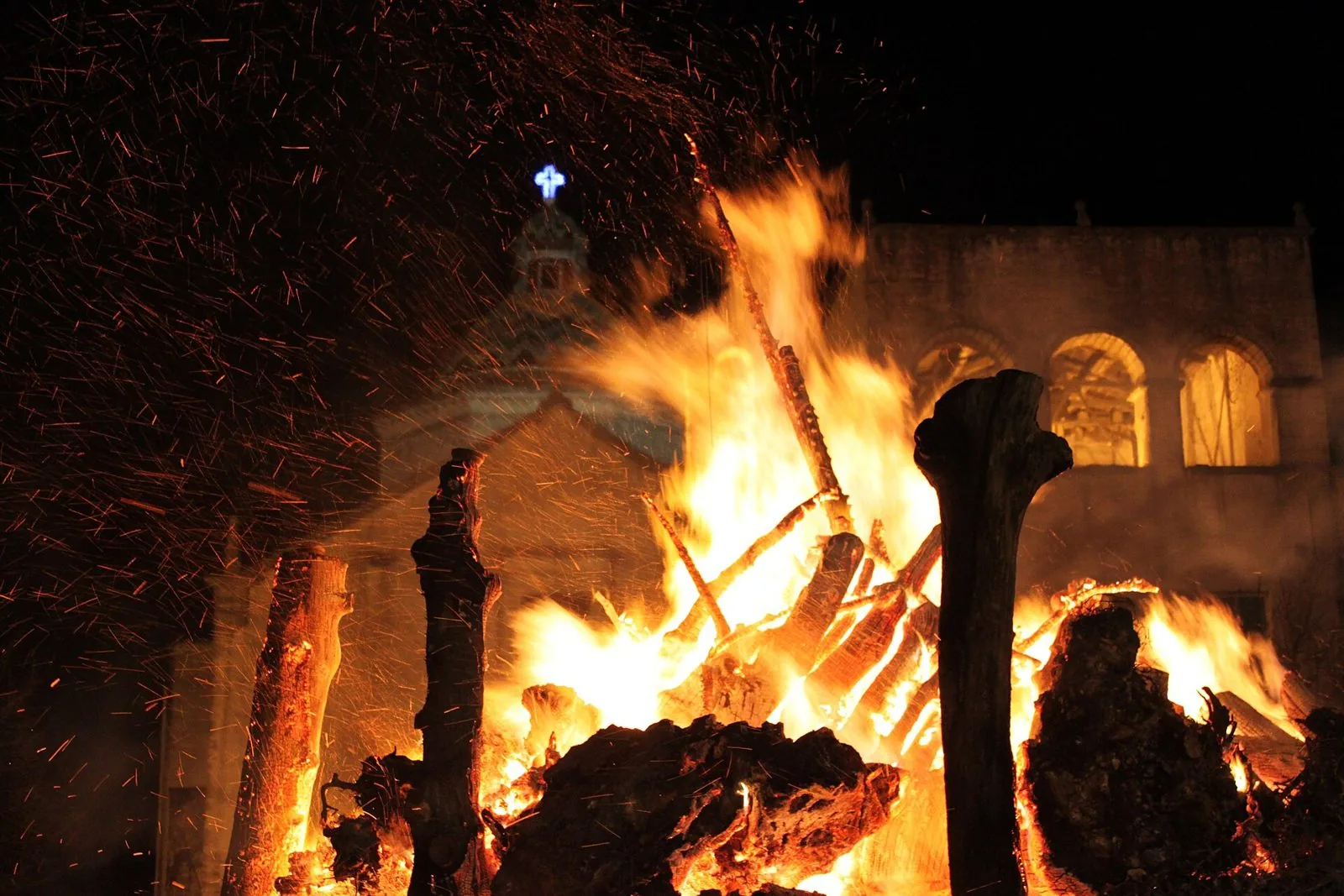 Un grande falò delle Fanove arde di notte con scintille e fiamme alte davanti alla facciata in pietra di un convento con arcate illuminate