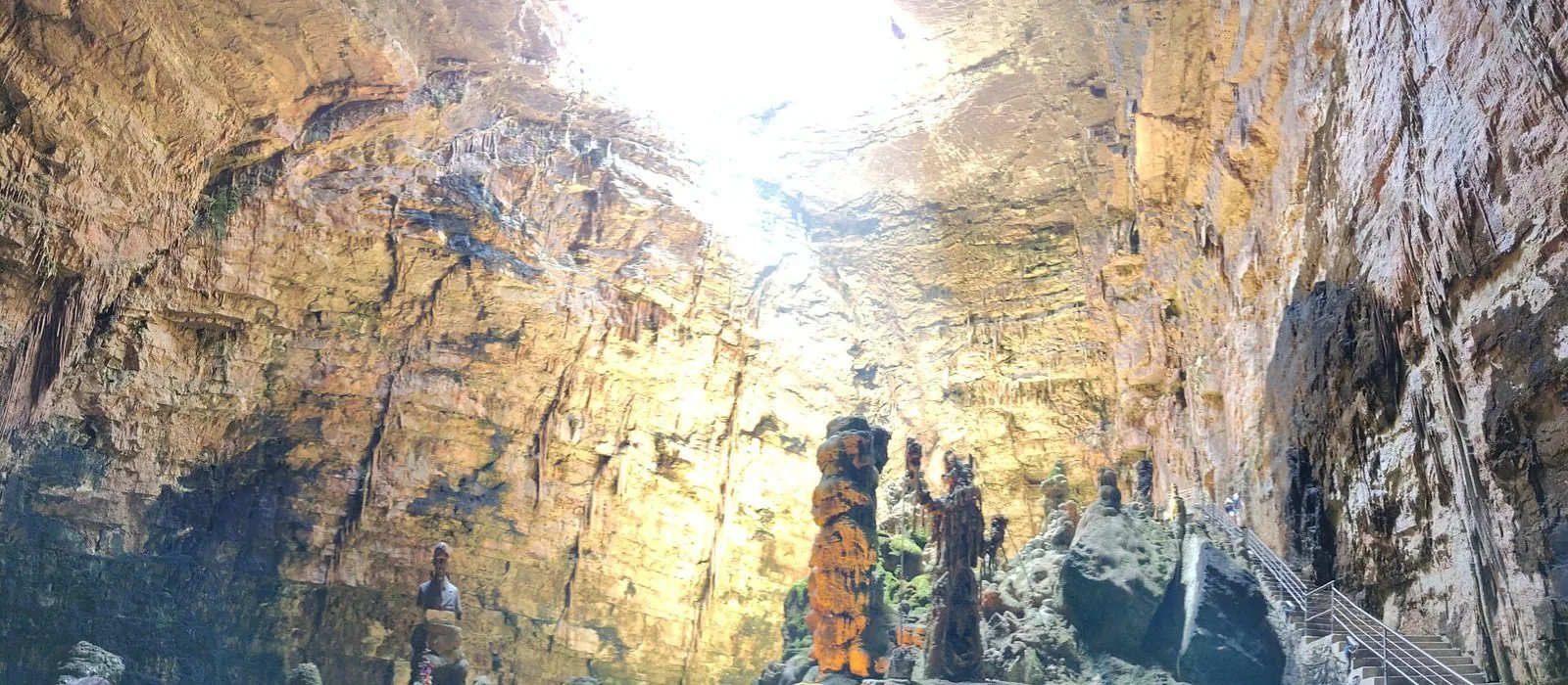 Interno della Grave delle Grotte di Castellana: alte pareti di roccia calcarea illuminate dalla luce che entra dall'apertura in alto, con stalagmiti e una scala sulla destra